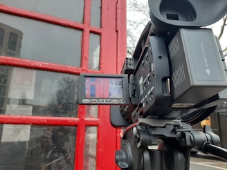 A digital video camera on a tripod is positioned in front of a red painted wooden structure with glass panes, likely a door or window. The camera's LCD screen displays the red structure, indicating that the camera is recording or preparing to record the scene. There are some reflections in the glass, and partial visibility of trees in the background.