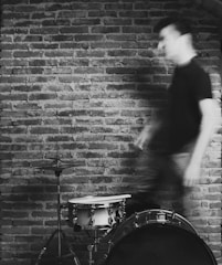 A blurred figure moves past a drum set against a textured brick wall, conveying a sense of motion and dynamism.