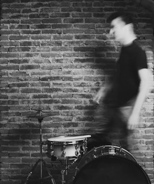 A blurred figure moves past a drum set against a textured brick wall, conveying a sense of motion and dynamism.