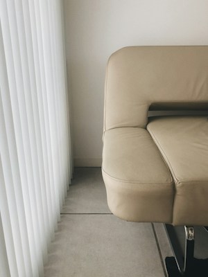 A modern beige leather sofa is positioned next to vertical white blinds, casting soft shadows on a tiled floor. The image captures a minimalist and clean setting with a focus on geometric lines and neutral tones.