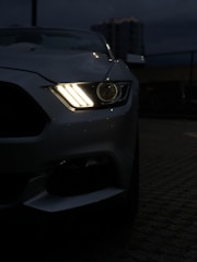 A close-up view of a car's front right headlight illuminated against a dim, urban night backdrop. The sleek design of the car, possibly a sports model, is partially visible with emphasis on the headlight's bright, stylish pattern.