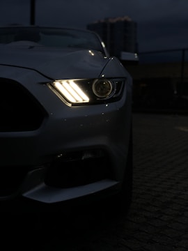 A close-up view of a car's front right headlight illuminated against a dim, urban night backdrop. The sleek design of the car, possibly a sports model, is partially visible with emphasis on the headlight's bright, stylish pattern.