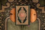A beautifully decorated book with intricate patterns and Arabic script, placed on a wooden book stand. The background features an ornate green and black patterned fabric, likely a prayer rug.