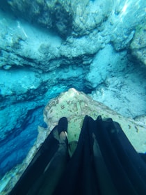 An underwater scene capturing clear, pristine blue water with a rocky terrain visible beneath. There is a person standing partially submerged, wearing a long dark skirt and black shoes, whose legs are visible from above.