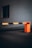 A brightly colored orange and black striped barrier gate positioned in front of a dark, neutral-toned background with a path leading up to it, alongside a wall with a small window.