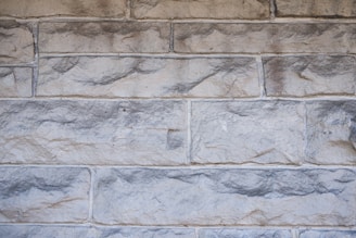 Rough stone wall with rectangular blocks, showing a variety of shades from light gray to beige. The stones have a textured, uneven surface with visible indentations and natural patterns.