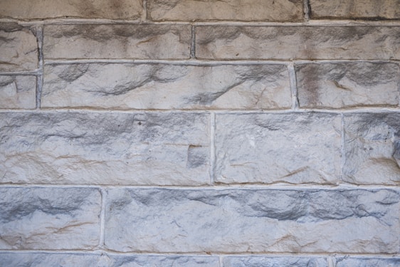 Rough stone wall with rectangular blocks, showing a variety of shades from light gray to beige. The stones have a textured, uneven surface with visible indentations and natural patterns.