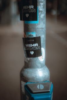 A pole is plastered with multiple stickers, featuring prominently the words 'MEHR DAVON' in bold with a heart symbol beneath. The pole itself has a metallic, weathered appearance, and there is a blue object attached to it at the bottom.