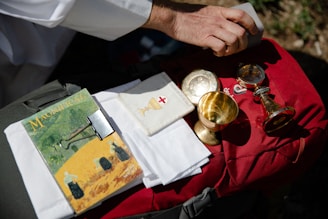 A hand holding a rectangular object over a red bag, which has a collection of religious items resting on it, including a magazine titled 'Magnificat' with a binder clip, white cloths, and several gold-colored chalices and containers.