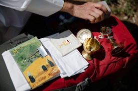 A hand holding a rectangular object over a red bag, which has a collection of religious items resting on it, including a magazine titled 'Magnificat' with a binder clip, white cloths, and several gold-colored chalices and containers.