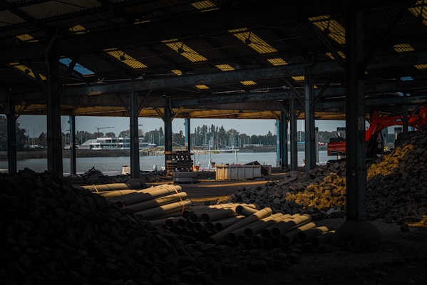 An industrial warehouse setting featuring construction materials such as pipes and large piles of rocks. Machinery, including an excavator, is visible, and the space is dimly lit. In the background, there is a view of a river and docked boats, with a partially cloudy sky adding to the moody atmosphere.