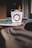 A Wok Box takeout container is centered on a glass table. The box is primarily white with red text and designs, and it features a metal handle. The background is blurry, with some indistinct objects and a cozy atmosphere.
