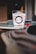 A Wok Box takeout container is centered on a glass table. The box is primarily white with red text and designs, and it features a metal handle. The background is blurry, with some indistinct objects and a cozy atmosphere.