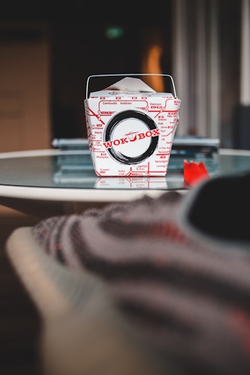 A Wok Box takeout container is centered on a glass table. The box is primarily white with red text and designs, and it features a metal handle. The background is blurry, with some indistinct objects and a cozy atmosphere.