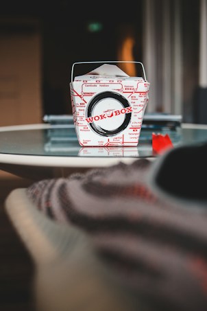 A Wok Box takeout container is centered on a glass table. The box is primarily white with red text and designs, and it features a metal handle. The background is blurry, with some indistinct objects and a cozy atmosphere.