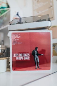An album cover prominently displays a red backdrop with a black and white image of a person. The text includes the album title 'Coming Home' along with a list of song titles.