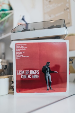 An album cover prominently displays a red backdrop with a black and white image of a person. The text includes the album title 'Coming Home' along with a list of song titles.