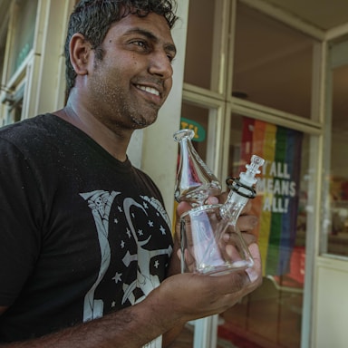 A person with short, curly hair and a beard is smiling while holding a clear glass object. They are standing outside in front of a building with a glass door. Behind them, there is a colorful flag with the text 'ALL MEANS ALL!'. The person is wearing a black t-shirt with a white graphic design.