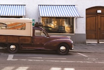 A vintage truck parked on a street in front of a building with two striped awnings. The truck has a canvas covering and wooden paneling with a retro sign printed on the side. The building features a large wooden door and a window displaying various items.