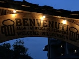 An illuminated wooden sign features the word 'Benvenuti' and the phrase 'Alla Fabbrica di Pedavena' beneath it, translating to 'Welcome' in multiple languages. The sign is adorned with icons of beer mugs on both sides, and several light bulbs illuminate it against a backdrop of a darkening sky.