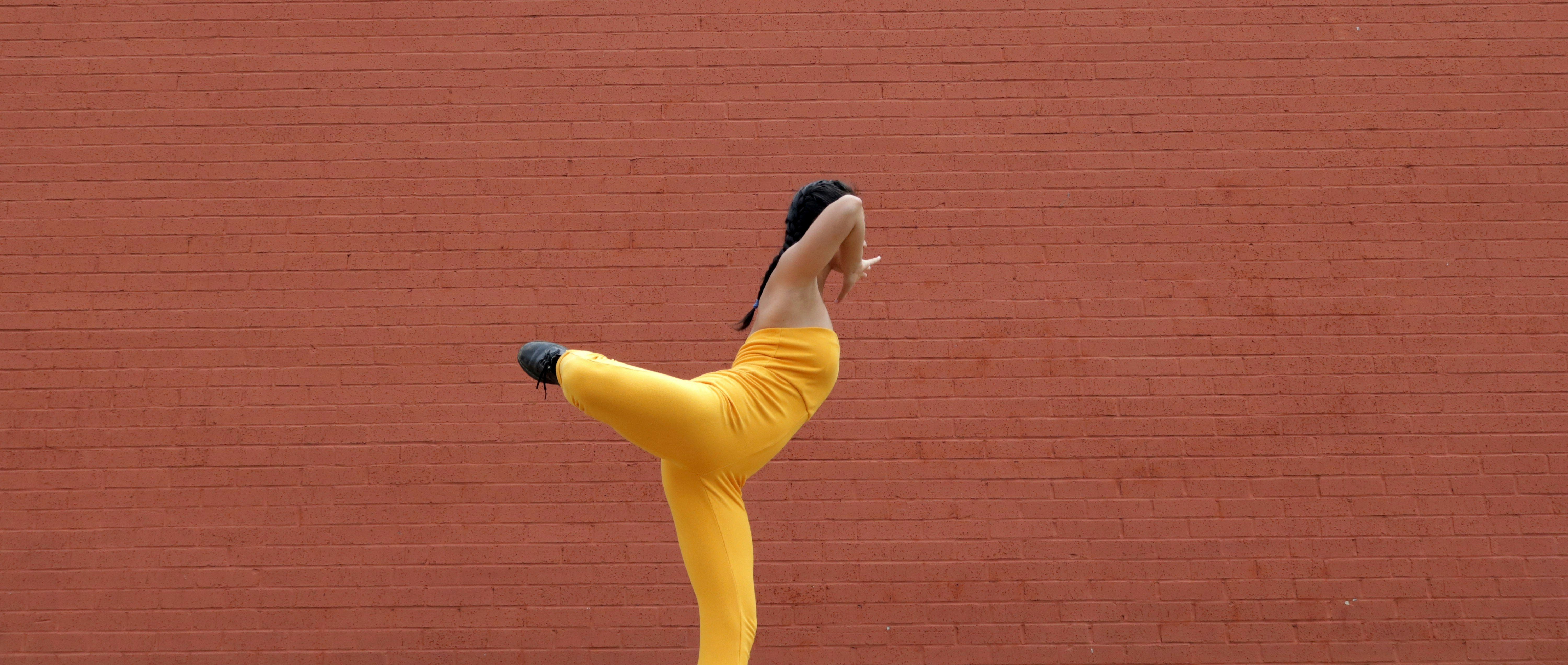 woman in yellow tank top and blue pants standing beside brown brick wall