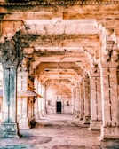 An architectural corridor with intricately carved pillars and arches, bathed in warm light. The design showcases traditional craftsmanship with detailed carvings on stone surfaces.