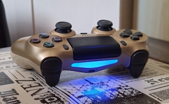 A gold-colored gaming controller with black thumbsticks and buttons rests on a surface covered with newspaper-style print. The light bar on the controller emits a bright blue glow.