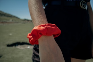 A close-up of a person's arm wearing a bright red scrunchie. The background is an outdoor setting with blurred greenery and blue sky, suggesting a sunny day. The person is wearing dark-colored shorts and a belt.