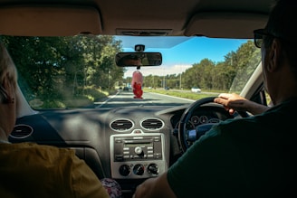 View from the inside of a car showing two people in the front seats. The driver is holding the steering wheel while driving on a tree-lined road with a clear blue sky. A red ornament hangs from the rearview mirror, and the dashboard displays the car's controls.