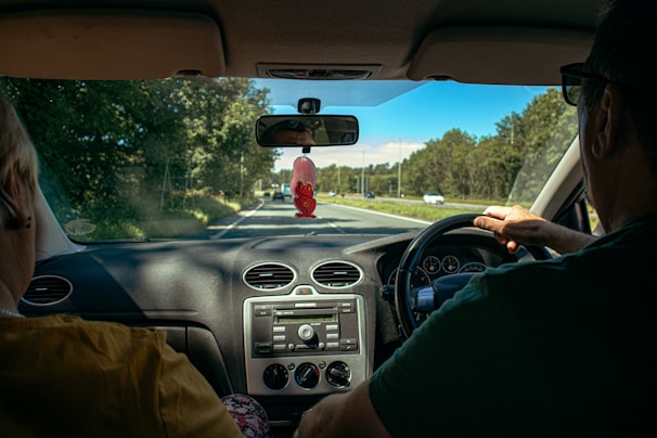 View from the inside of a car showing two people in the front seats. The driver is holding the steering wheel while driving on a tree-lined road with a clear blue sky. A red ornament hangs from the rearview mirror, and the dashboard displays the car's controls.