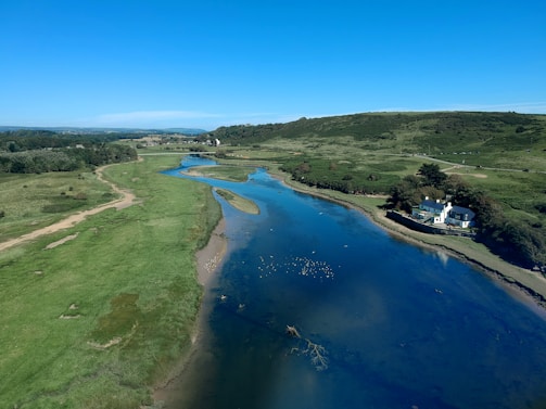A calm river flows through a lush, green landscape under a clear blue sky. A house is situated on the right bank, surrounded by trees. The river reflects the blue of the sky and is dotted with small patches of vegetation. Rolling hills stretch into the background.
