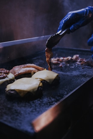 A person wearing blue gloves is using tongs to flip and cook bacon on a hot, flat grill. Next to the bacon, there are several burger patties topped with melting slices of cheese, indicating the preparation of cheeseburgers.