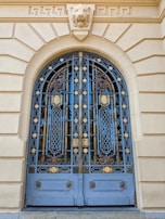 Ornate wrought iron doors featuring intricate geometric and floral patterns, set within a stone archway. Above the doors is a stone lion sculpture, adding a majestic touch to the architecture.