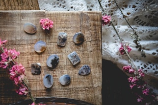 A collection of polished stones with intricate, dendritic patterns is arranged on a rustic wooden surface. Delicate pink flowers are scattered across the scene, adding a natural and artistic touch. The background includes a textured, lace fabric enhancing the overall vintage and bohemian vibe.