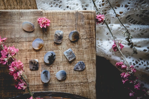 A collection of polished stones with intricate, dendritic patterns is arranged on a rustic wooden surface. Delicate pink flowers are scattered across the scene, adding a natural and artistic touch. The background includes a textured, lace fabric enhancing the overall vintage and bohemian vibe.