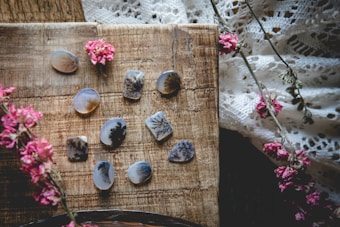 A collection of polished stones with intricate, dendritic patterns is arranged on a rustic wooden surface. Delicate pink flowers are scattered across the scene, adding a natural and artistic touch. The background includes a textured, lace fabric enhancing the overall vintage and bohemian vibe.