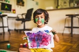 A young child with paint on their face and hands holds up a colorful painting. The artwork consists of vibrant splashes of purple, pink, blue, and green. The background shows a home setting with wooden floors, a desk with a computer, and chairs.