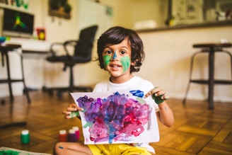 A young child with paint on their face and hands holds up a colorful painting. The artwork consists of vibrant splashes of purple, pink, blue, and green. The background shows a home setting with wooden floors, a desk with a computer, and chairs.