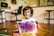 A young child with paint on their face and hands holds up a colorful painting. The artwork consists of vibrant splashes of purple, pink, blue, and green. The background shows a home setting with wooden floors, a desk with a computer, and chairs.