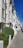 An elegant row of white, multi-story townhouses with large windows, aligned perfectly along a clean, paved sidewalk. Manicured green hedges are neatly placed in front of each building, and the sky is clear and blue, giving the street a bright and inviting appearance.
