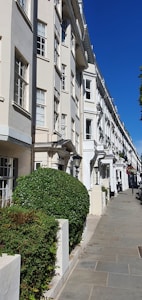 An elegant row of white, multi-story townhouses with large windows, aligned perfectly along a clean, paved sidewalk. Manicured green hedges are neatly placed in front of each building, and the sky is clear and blue, giving the street a bright and inviting appearance.