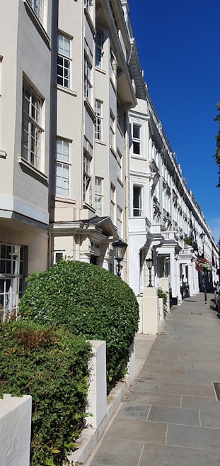 An elegant row of white, multi-story townhouses with large windows, aligned perfectly along a clean, paved sidewalk. Manicured green hedges are neatly placed in front of each building, and the sky is clear and blue, giving the street a bright and inviting appearance.