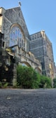 A large stone church with tall arched windows featuring intricate designs. The building is surrounded by lush greenery and shrubs. The architecture is gothic-inspired, with pointed arches and detailed carvings.