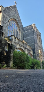 A large stone church with tall arched windows featuring intricate designs. The building is surrounded by lush greenery and shrubs. The architecture is gothic-inspired, with pointed arches and detailed carvings.