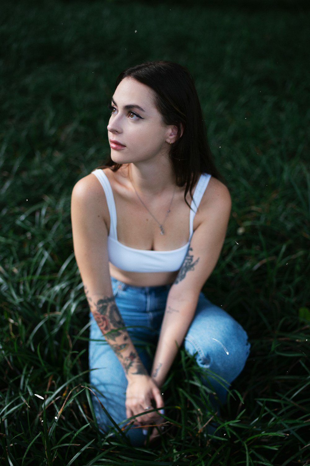 woman in white tank top and blue denim jeans sitting on green grass field