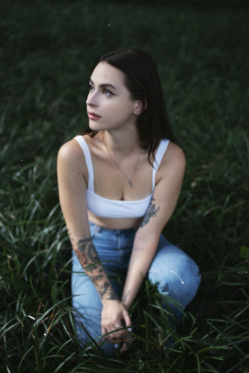 woman in white tank top and blue denim jeans sitting on green grass field