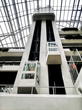 A modern architectural structure featuring large glass elevators within a building atrium. The ceiling consists of an intricate network of glass and metal, allowing natural light to flood the area. The elevators are sleek and enclosed in glass, with visible cables and structural elements. Potted greenery adorns each level, adding a touch of nature to the contemporary design.