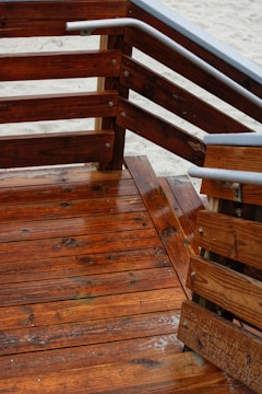 Wooden deck with a series of stairs and railings. The wood is richly colored and appears freshly varnished, with a glossy finish. The grain of the wood is clearly visible, and there are metal accents on the railings. The sandy ground is visible in the background.