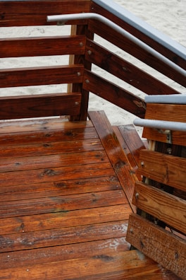 Wooden deck with a series of stairs and railings. The wood is richly colored and appears freshly varnished, with a glossy finish. The grain of the wood is clearly visible, and there are metal accents on the railings. The sandy ground is visible in the background.