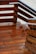 Wooden deck with a series of stairs and railings. The wood is richly colored and appears freshly varnished, with a glossy finish. The grain of the wood is clearly visible, and there are metal accents on the railings. The sandy ground is visible in the background.